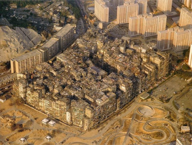 Kowloon Walled City. A large solid block of ramshackle buildings varying in height, with many taller buildings and some mountains in the background. – Author: Ian Lambot – CC BY-SA 4.0
