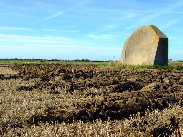 The farmland has been ploughed around the mirror. The rusty pole in front of the mirror had a microphone attached to it when listening for aircraft in WWI. Author: John Poyser CC BY SA2.0