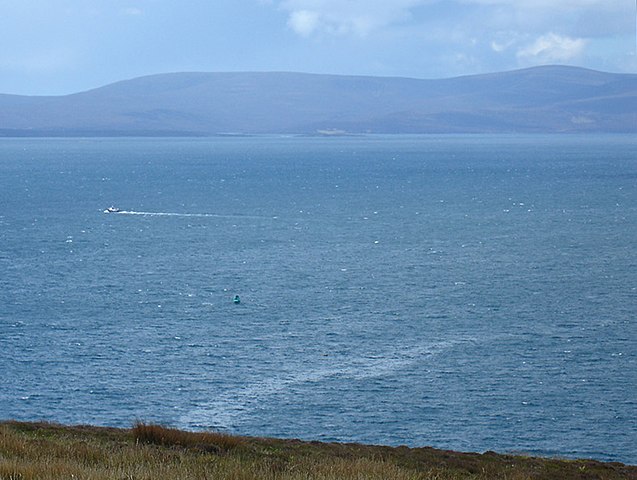Scapa Flow from Gaitnip cliffs, showing the wreck site of HMS Royal Oak marked by a green buoy. An oil slick is visible rising to the surface after almost 80 years. Author: BillC CC BY-SA 3.0