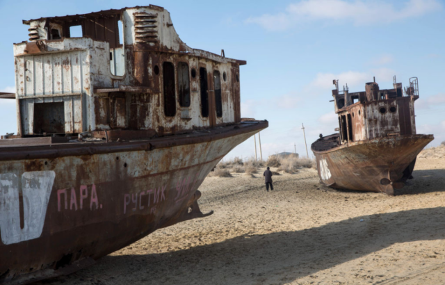 Man standing between two rusty ships in the middle of the desert