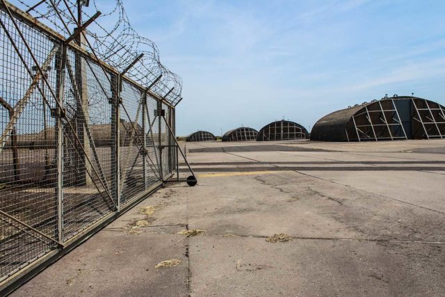 Concrete ground with a barbed wire fence to the left and an airplane hanger to the right