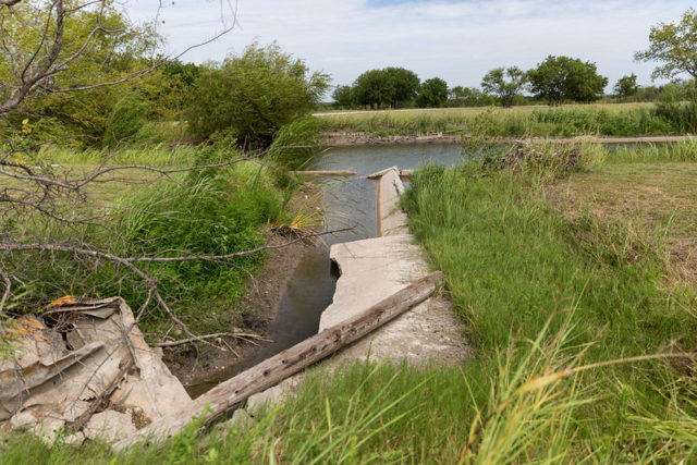 Broken cement surrounded by grass, leading to a storm shelter filled with water