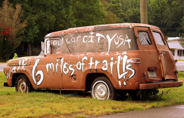 Rusty car with white paint that reads "Old Car City, USA / 6 miles of trails"