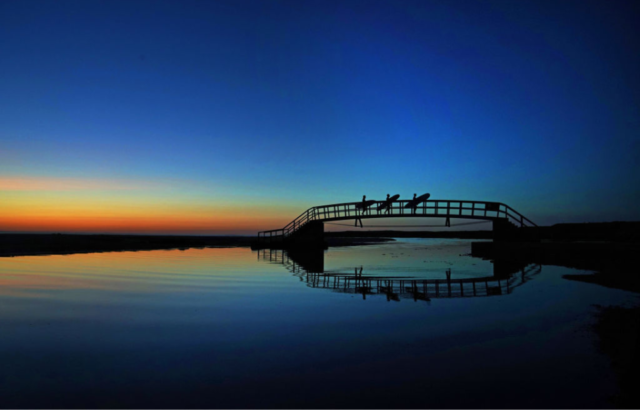 Three people crossing Belhaven Bridge while carrying surfboards