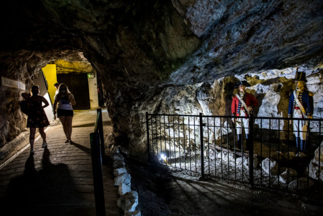 People walking through a rock tunnel with two soldier statues located behind a fence on their right.