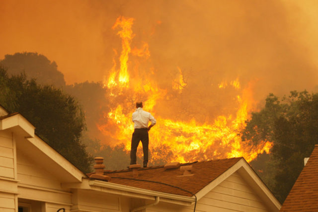 Man standing on the roof of a house looking at a large forest fire.