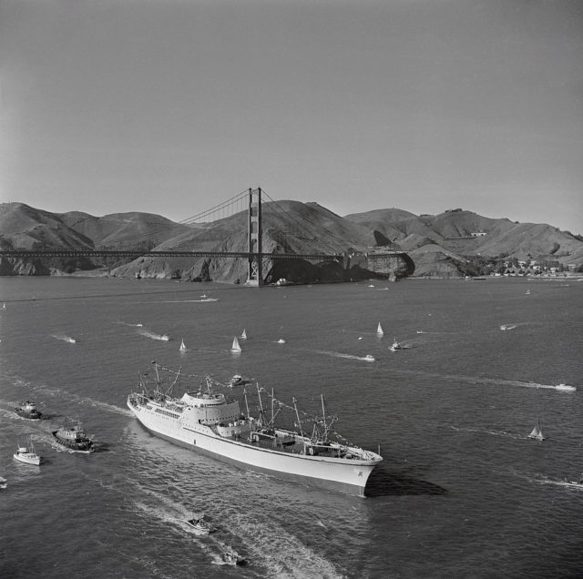 Black and white photo of a group of boats traveling on the water with the Golden Gate Bridge in the background. 