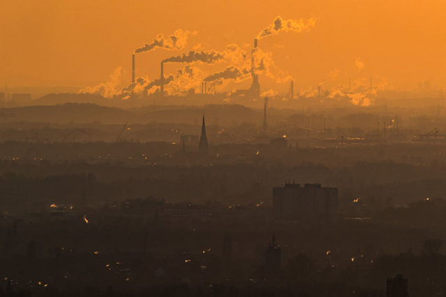 Smoke coming out of smoke stacks in front of an orange sky.