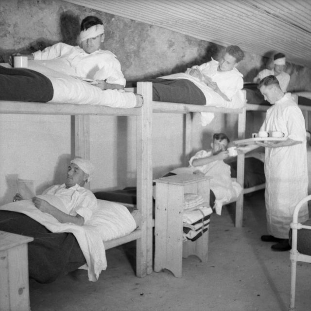 Wounded soldiers lying on bunk beds in a room made of rock while a doctors checks on them.