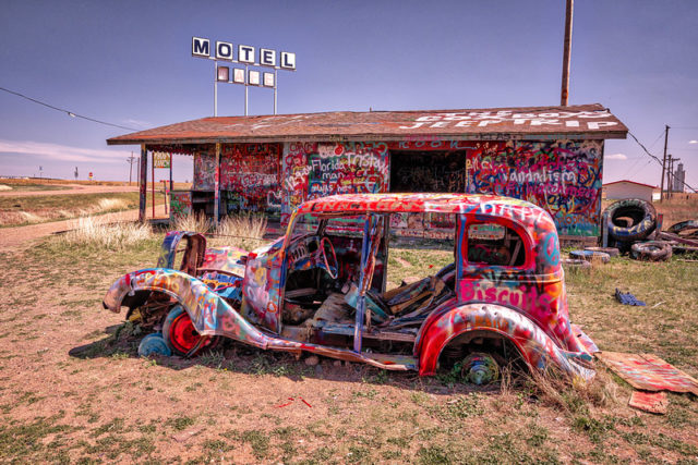 Old car spray painted and without windows and doors sitting in front of a spray painted building.