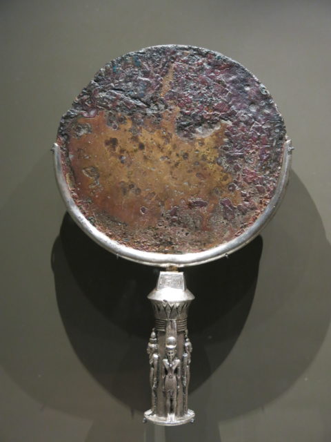 Tarnished and rusting mirror with a silver handle detailed with humanoids standing around the handle.