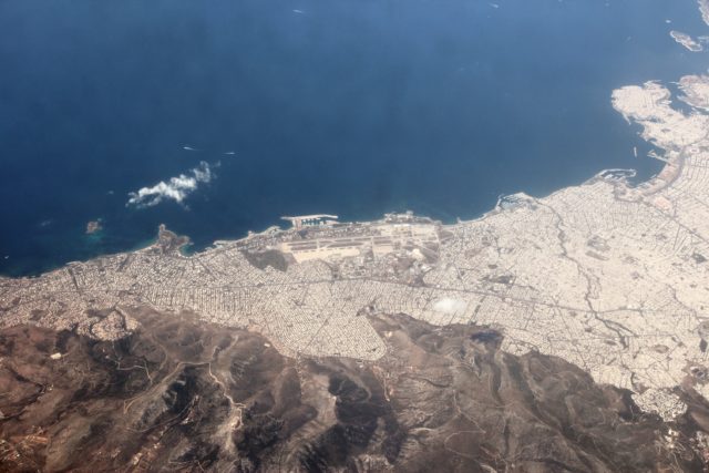 Aerial view of the old Ellinikon International Airport on the coast with sprawling cities around it, and mountain ranges in the background.