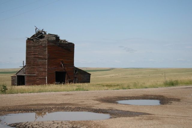 Abandoned wooden building breaking apart at the top with a road and puddles in the foreground.