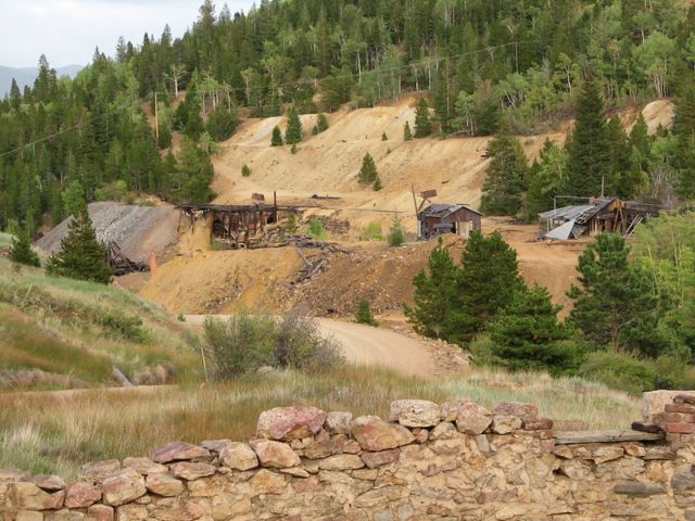 Abandoned buildings falling apart on a sandy hill with trees in the background and a stone wall in the foreground. 