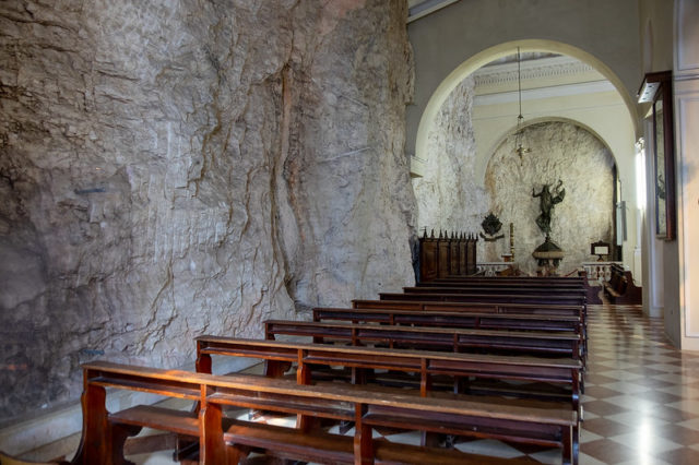 Interior of a chapel with one wall made entirely out of a rock face, with pews running down the middle of the room towards the alter. 