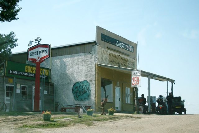 Side view of the yellow gas station with a red sign reading "Ghost Town" beside it and people on motorcycles filling up with gas.