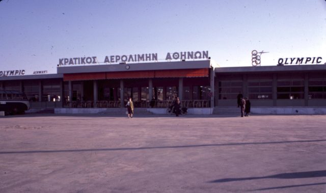 Airport terminal with a sign in Greek letters above it and people milling around outside it.