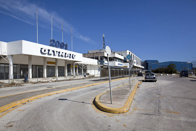 Entrance to Ellinikon International Airport with a white facade that reads "2001 Olympic" with a chain fence around it.