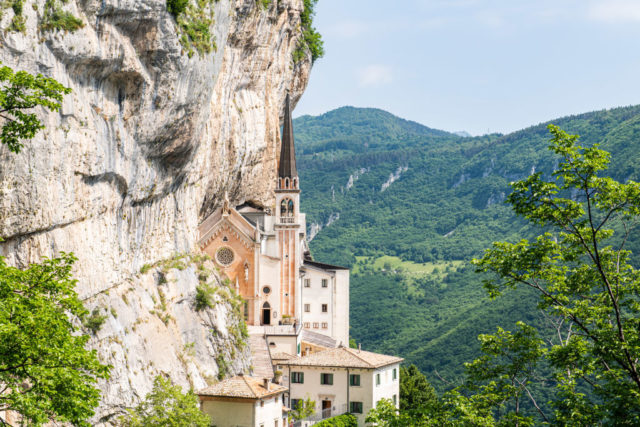 Distanced view of Santuario Madonna della Corona in the rock face vibrant green trees covering the mountain in the background.