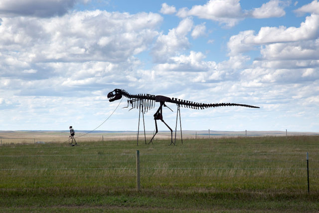 Statue of a dinosaur being led by a person through an open grass field.