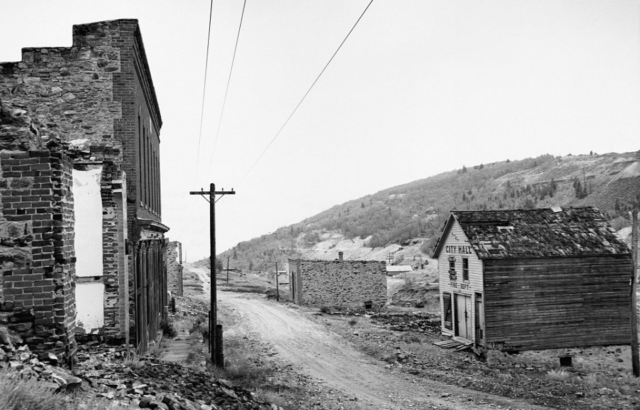 Black and white image looking down a dirt road with abandoned and destroyed brick buildings on either side.