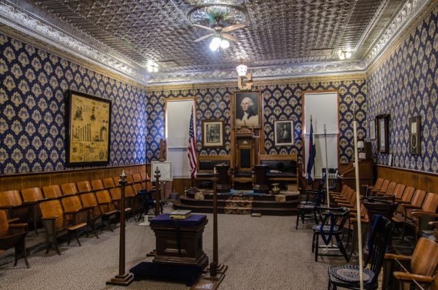 Room of the Masonic Lodge lined with wooden chairs with a raised dais with photos behind it. 