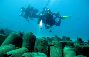 Two divers underwater with flashlight looking down at multiple clay jars scattered on the ground.