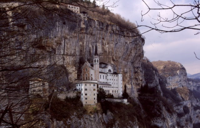 Distanced view of Santuario Madonna della Corona, an old white church, built on the side of a cliff.