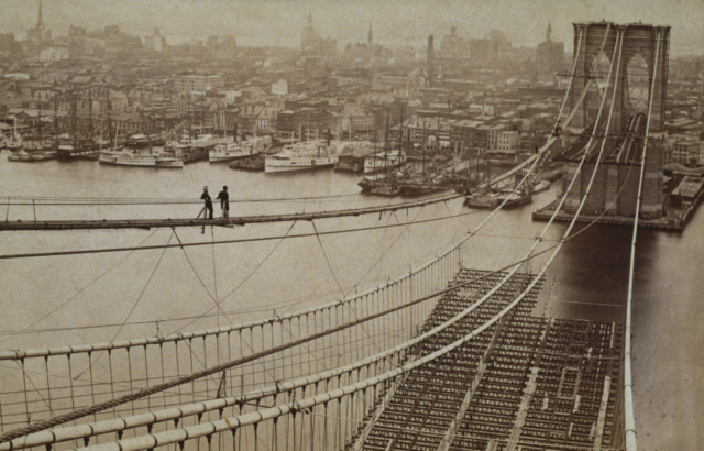 Two men stand over the incomplete Brooklyn Bridge during construction
