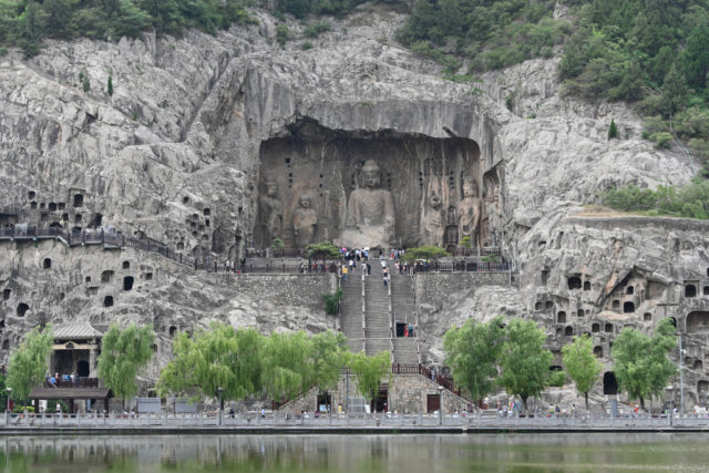 Distanced shot of a large Buddha statue carved into a cliff side with stairs leading up to it and small windows carved out in the surrounding rock.