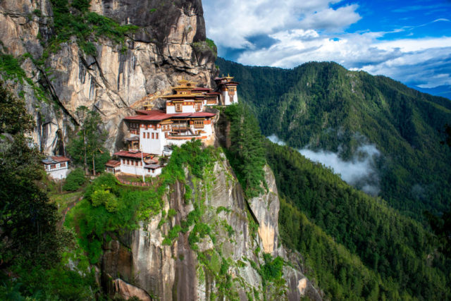 Ornate monastery with white walls and a red roof situated on a heavily treed mountain side.