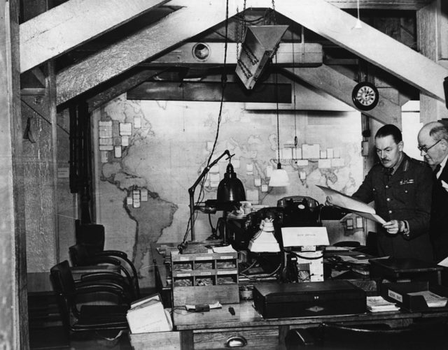 Government officials looking over a document in a cluttered room in the Cabinet War Rooms