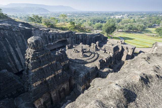 Ellora Caves, detailed statues and buildings carved out of dark rock with trees in the distance.
