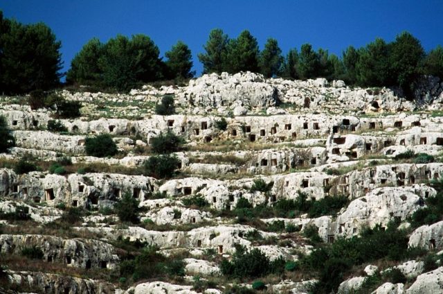 Necropolis of Pantalica, tiered rocks with windows cut in them and trees at the top of the hill.