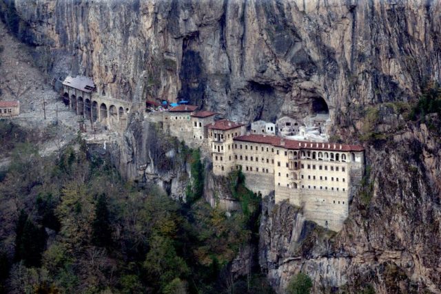 Aerial view of the Sumela Monastery, a stone building with multiple floors and a red roof, build into the side of a tall cliff.