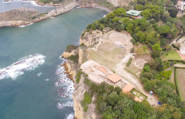 Aerial view of ancient Roman buildings surrounded by green trees on one side, and a body of water on the other.