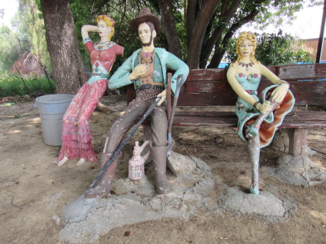 Sculpture of two women and a man sitting on a bench in old Western attire.