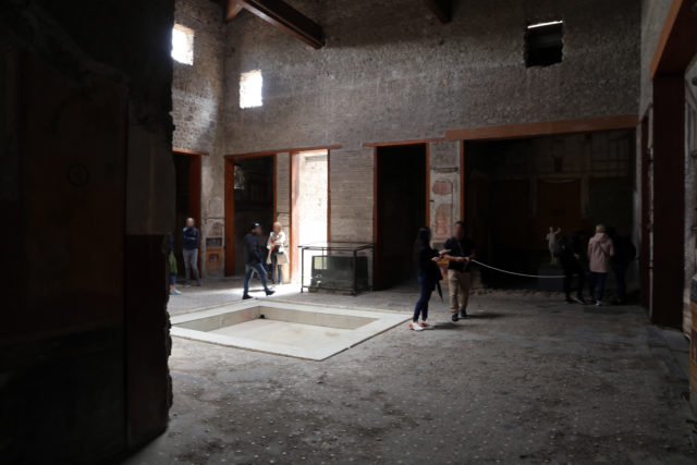People walking inside a dark Roman room, many doorways leading off of the room.
