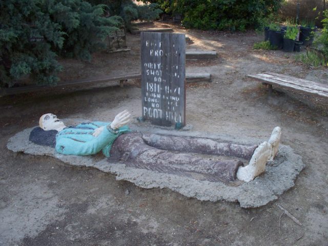 Sculpture of a man laying on the ground, a tombstone with faded words behind him