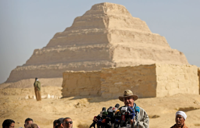 Man standing behind microphones on a podium with a large step pyramid in the background.