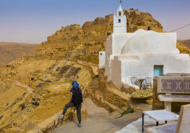 A person standing beside a white mosque, a hilltop village behind it