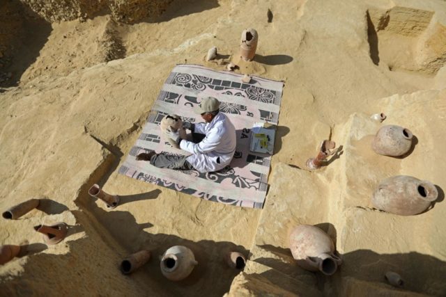 Man sitting on a rug in the sand with old Egyptian pottery surrounding him.