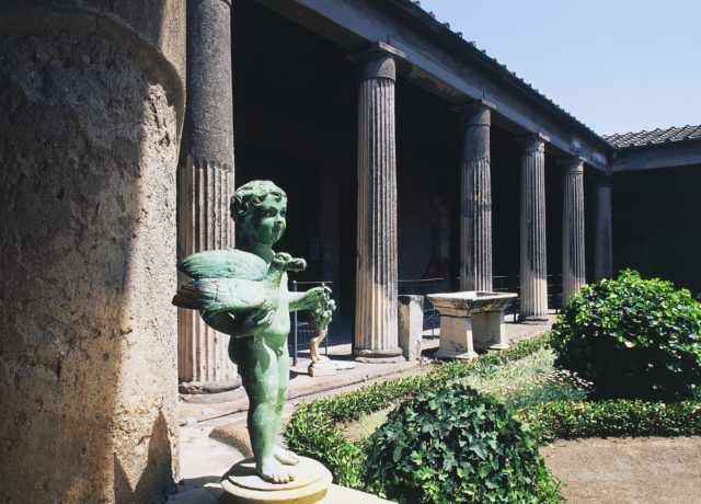 A small green cupid statue in front of pillars holding up a roof outside of a garden.