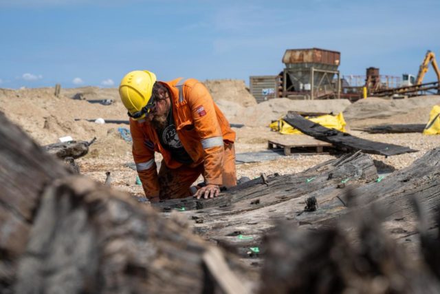 Archaeologist in an orange jumpsuit with yellow hard hat inspecting a wooden ship hull.