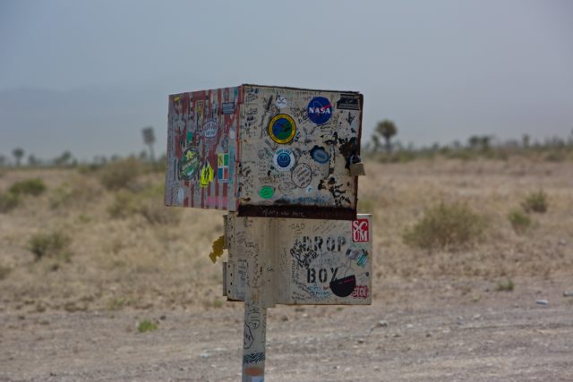 A mailbox containing two boxes atop one another, covered in stickers in the desert.