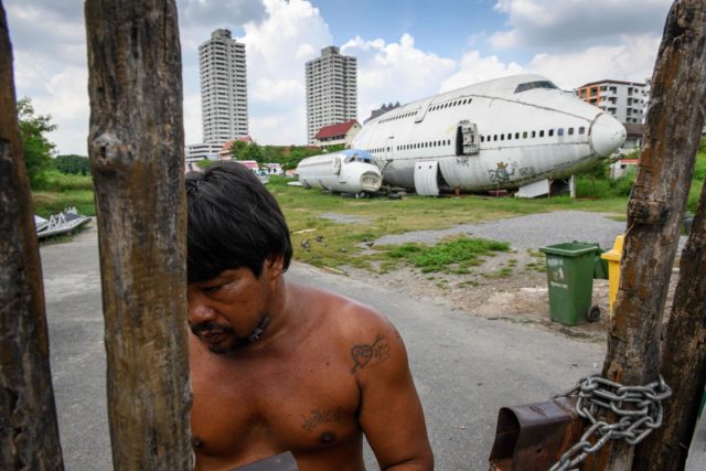 A man opens and wood gate with an abandoned airplane behind him. 