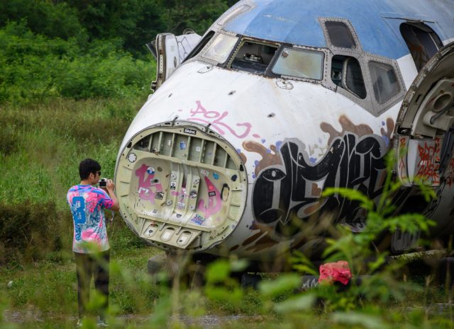 A tourist stands in front of an abandoned and graffitied airplane sitting on a lot.