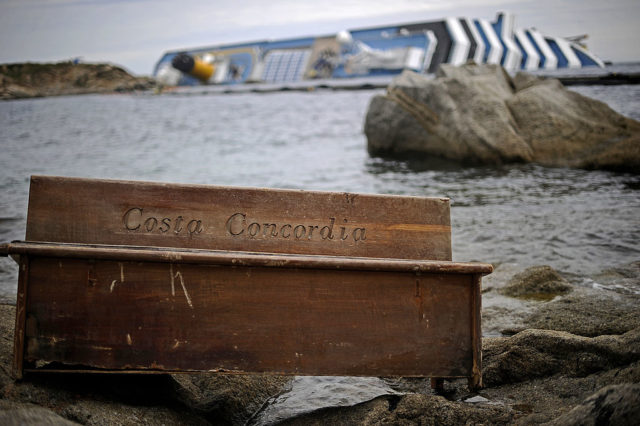 Bench with the etching 'Costa Concordia' washed up on the beach, with the ship in the background.