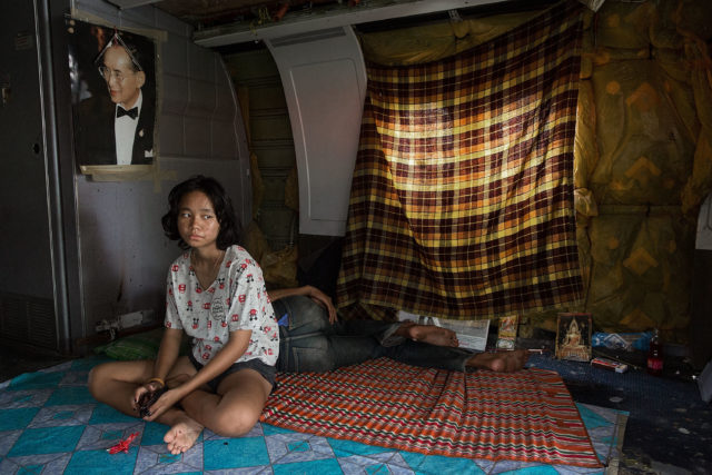 A girl sitting on the ground with blankets on the floor, a curtain over a window, and a poster of a man on the wall. 