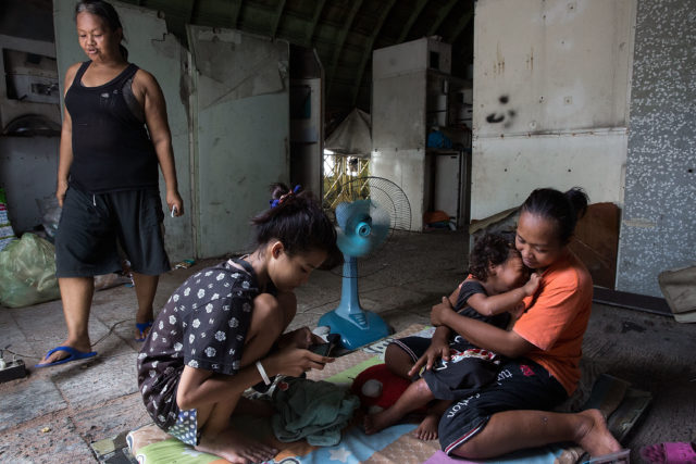 A family sits on the ground of a room, a woman walking past.